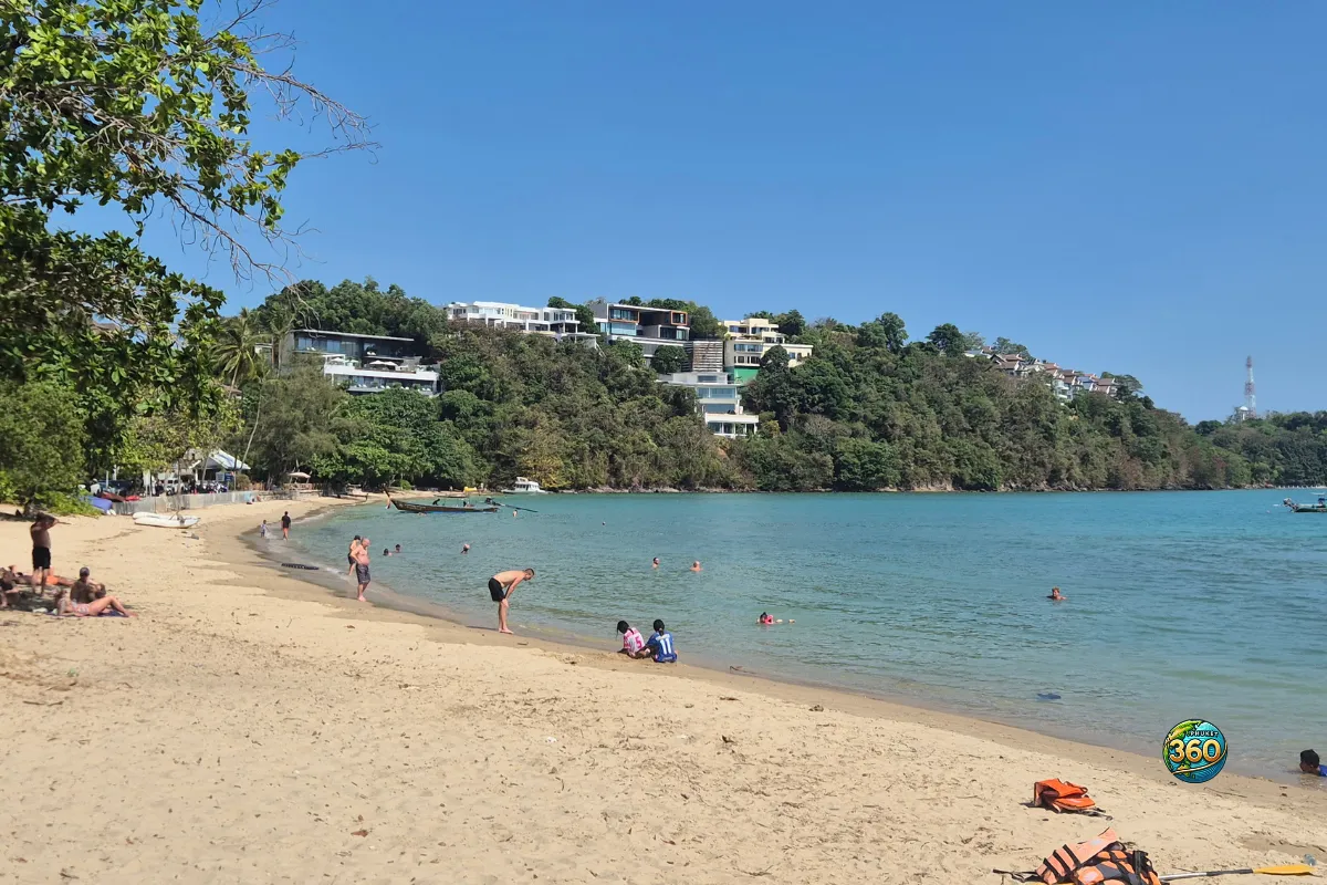 Vue sur la baie d’Ao Yon Beach à Phuket, avec une plage de sable, des baigneurs, une mer calme et des villas sur la colline
