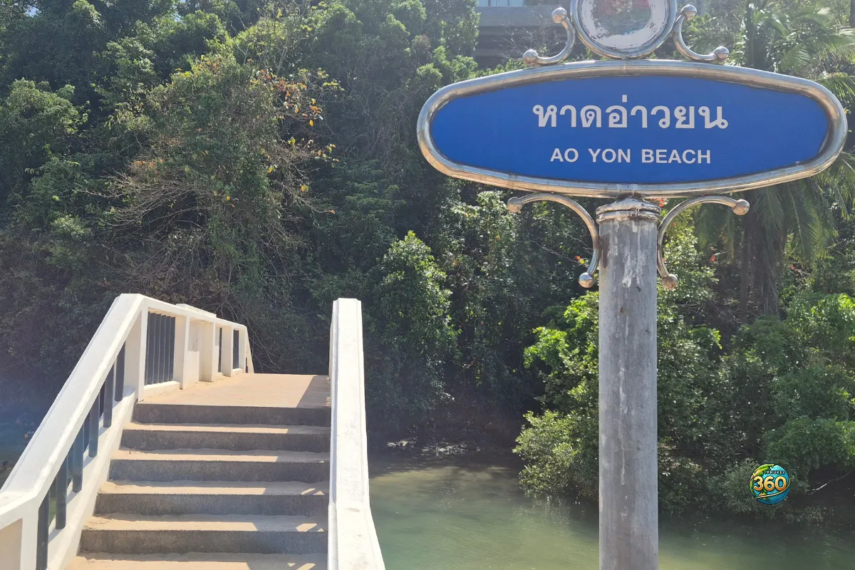 Panneau Ao Yon Beach à Phuket à côté d’un petit pont, avec de la végétation tropicale autour