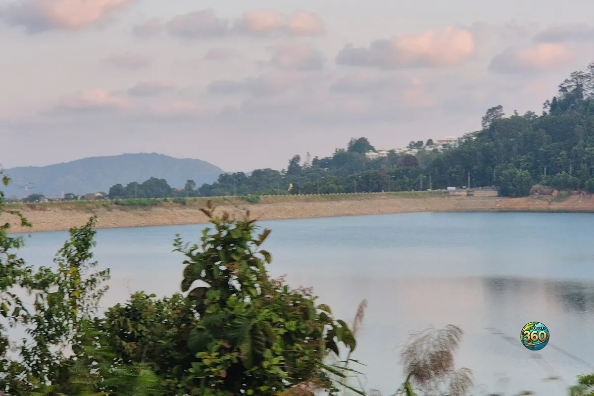 Vue sur le lac de Chalong Dam Reservoir à Phuket avec végétation au premier plan, digue rocheuse et ciel pastel