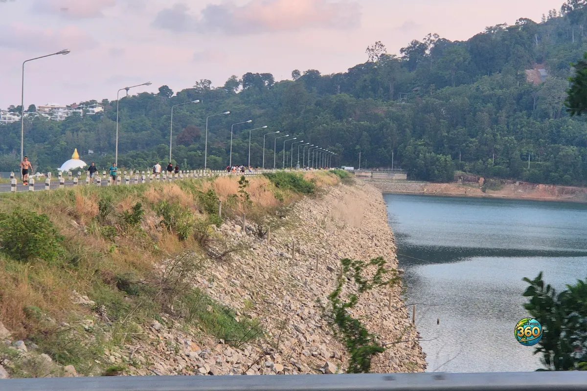 Route sur la digue de Chalong Dam Reservoir à Phuket avec lac, collines boisées et promeneurs au coucher du jour