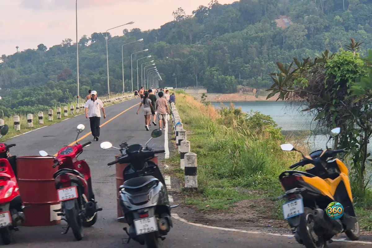 Scooters garés au bord de la route de Chalong Dam Reservoir à Phuket avec promeneurs, lac et colline en arrière-plan