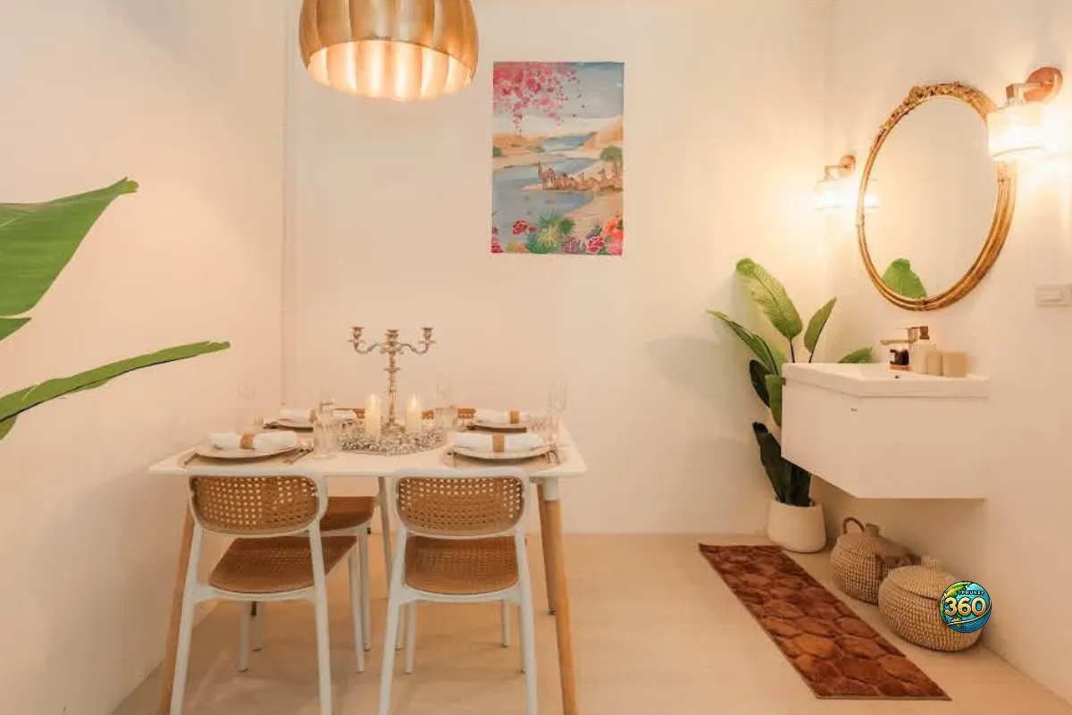 Coin repas décoré d’une chambre à louer à Phuket avec table dressée, lavabo mural, miroir rond, plantes et lumière chaude