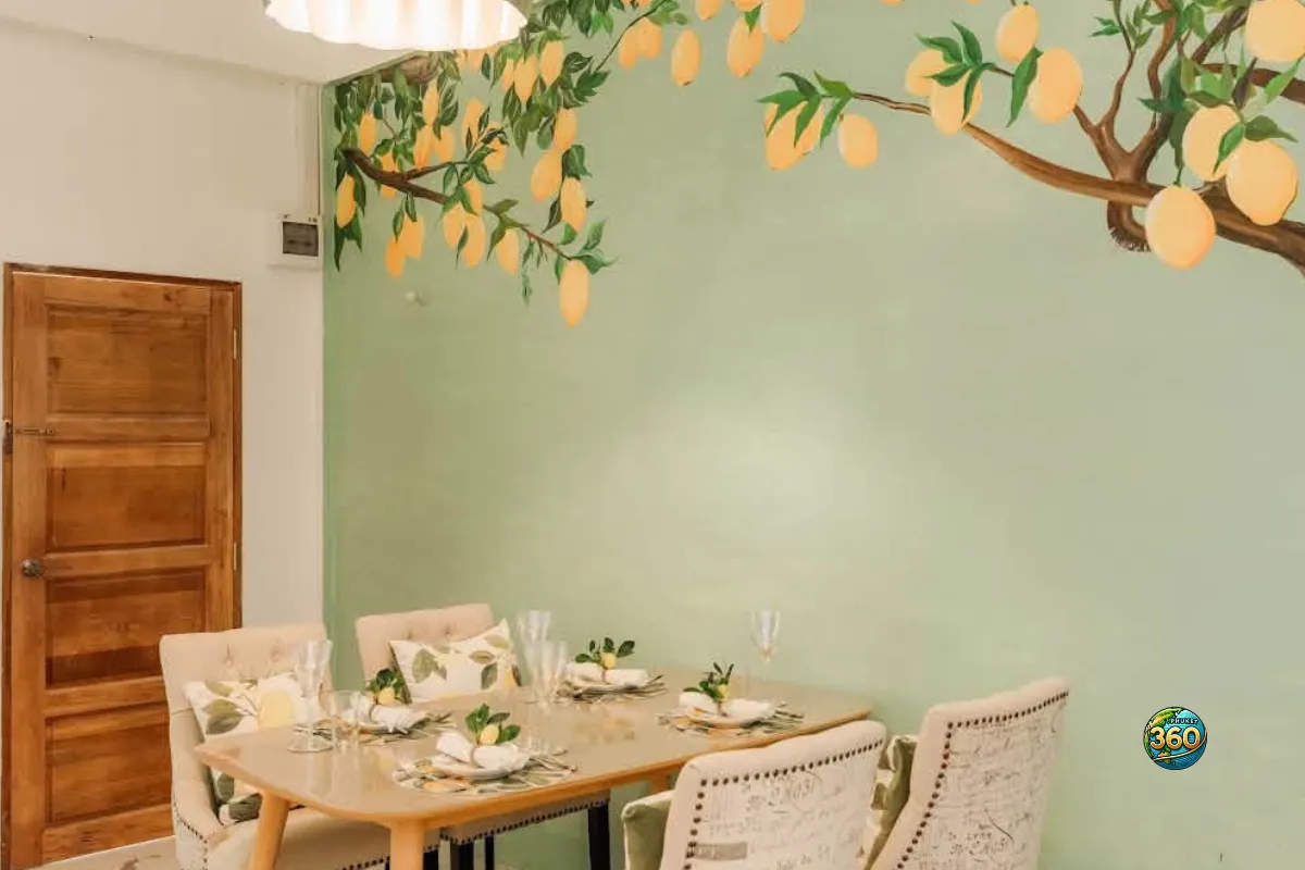 Coin repas d’une chambre à louer à Phuket avec table dressée, chaises blanches, porte en bois et mur décoré de citrons