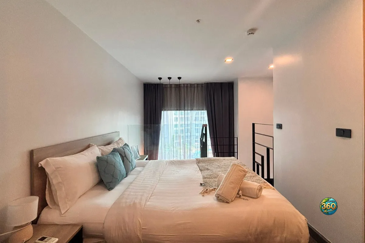 Chambre en mezzanine du duplex à Utopia Nai Harn, Phuket, avec lit double, linge de lit clair, garde-corps vitré et vue vers la baie vitrée
