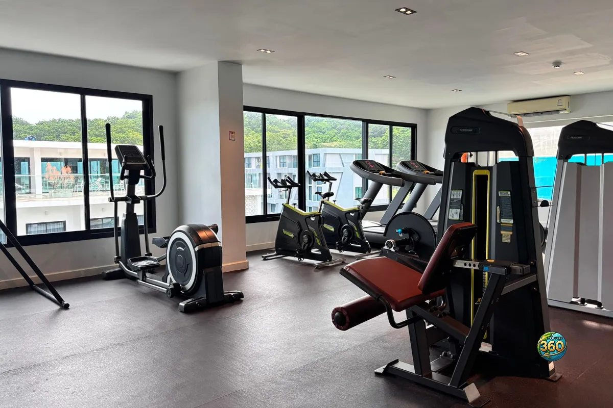 Salle de sport de la résidence Utopia Nai Harn à Phuket avec vélos, tapis de course, machines de musculation et grandes fenêtres