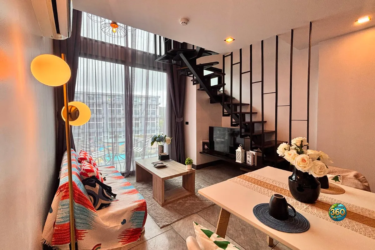 Espace salon et salle à manger du duplex à Utopia Nai Harn, Phuket, avec grande baie vitrée, escalier noir et cuisine ouverte