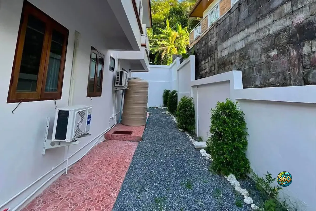 Allée latérale de la maison 3 chambres à louer à Bang Tao, Phuket, avec gravier, plantes, unités de climatisation et réservoir d’eau