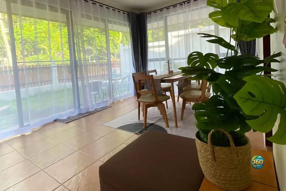 Espace repas lumineux de la maison 3 chambres à louer à Bang Tao, Phuket, avec table en bois, grandes baies vitrées, rideaux et plante intérieure