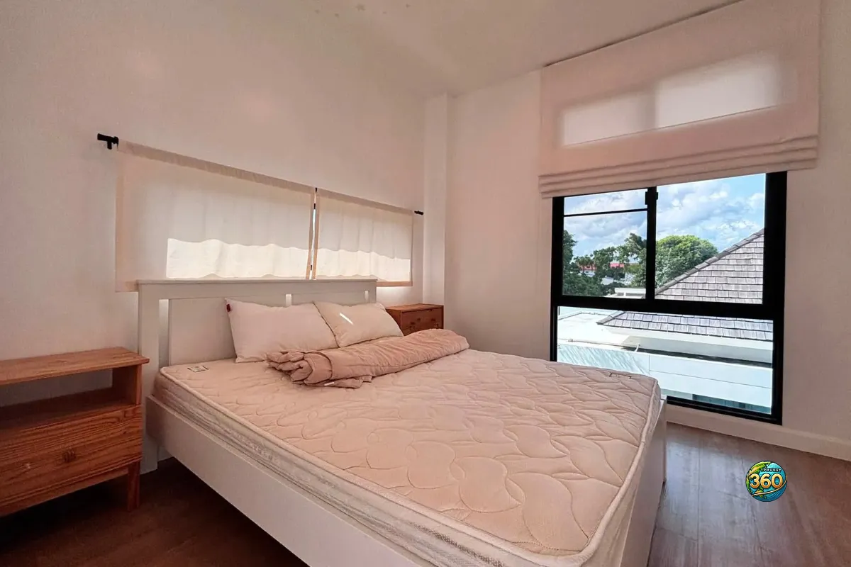 Chambre meublée de la maison Mono Plus Palai à Phuket avec lit double, tables de chevet, grande fenêtre et sol imitation bois