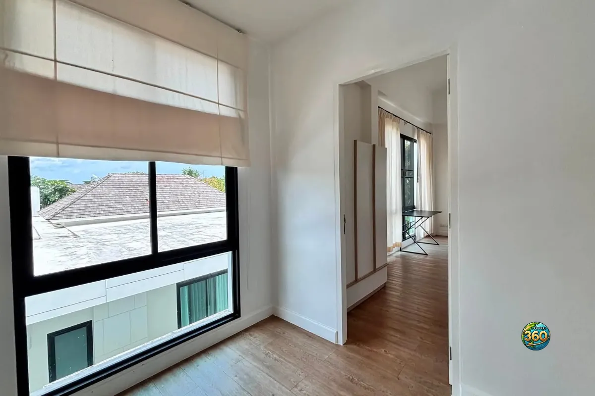 Chambre vide de la maison Mono Plus Palai à Phuket avec grande fenêtre, sol imitation bois et vue vers une autre pièce meublée