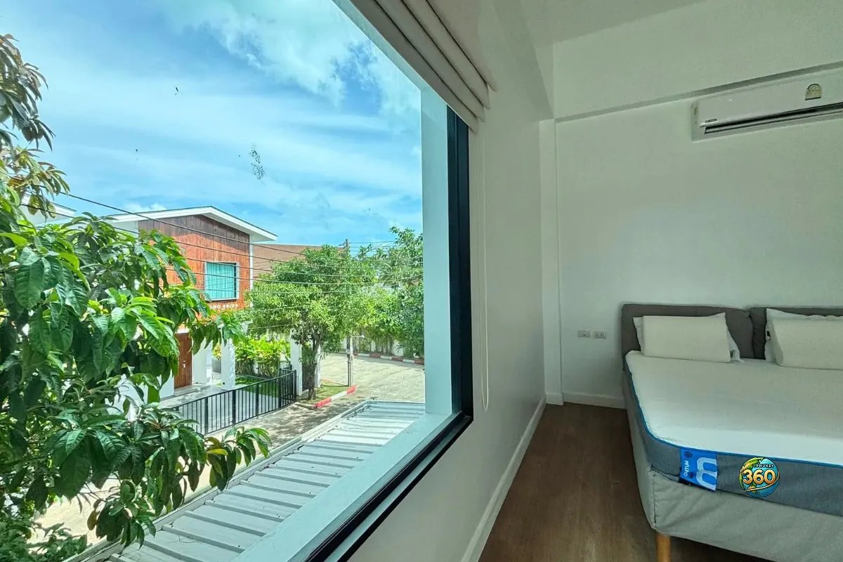 Chambre lumineuse de la maison Mono Plus Palai à Phuket avec lit, climatisation, grande fenêtre et vue sur l’extérieur de la résidence