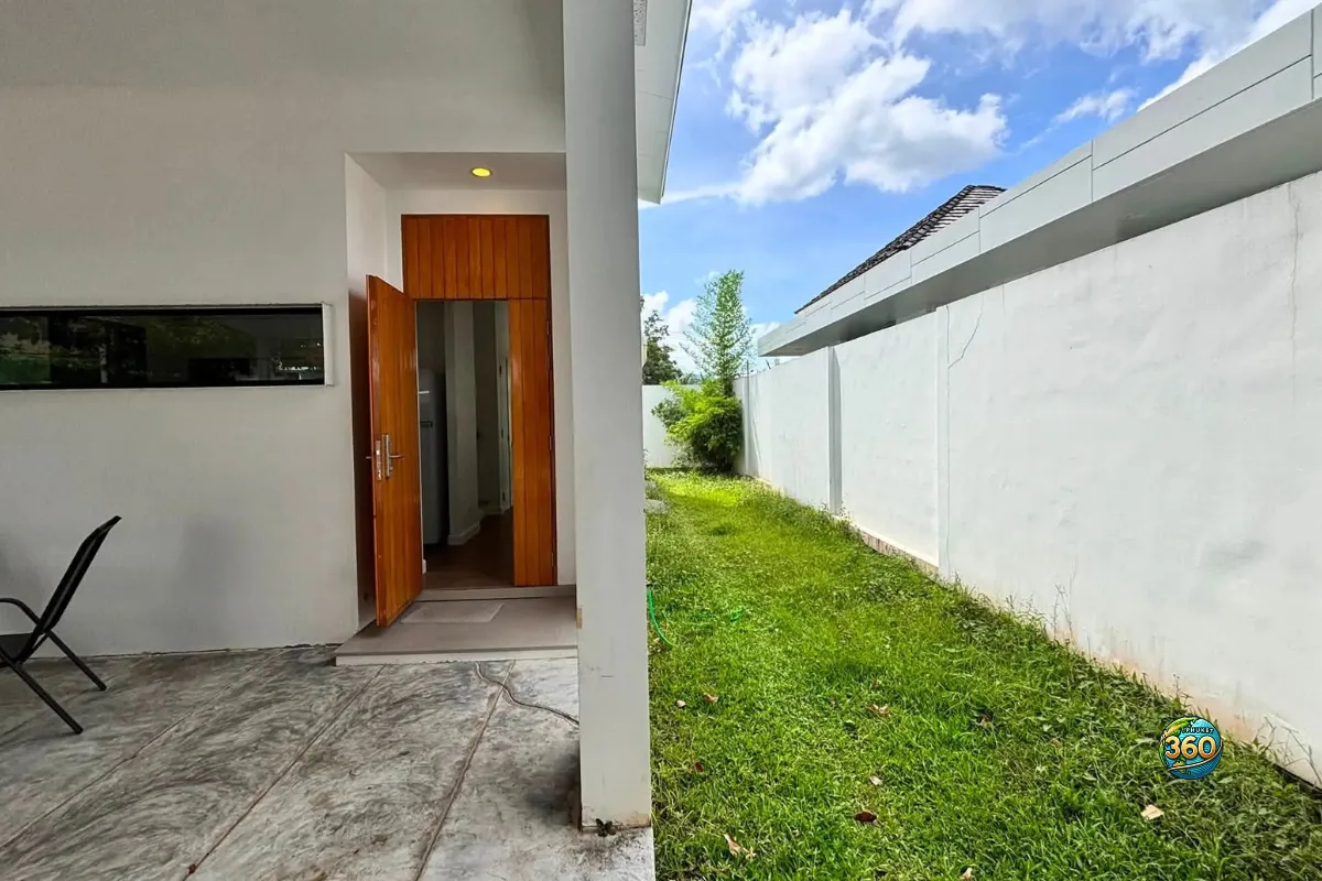 Jardin latéral de la maison Mono Plus Palai à Phuket avec entrée ouverte, terrasse couverte et mur de clôture blanc