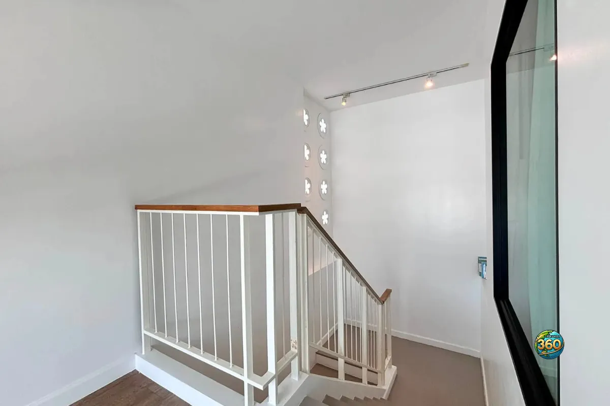 Palier à l’étage de la maison Mono Plus Palai à Phuket avec escalier, rambarde blanche, mur lumineux et grande fenêtre