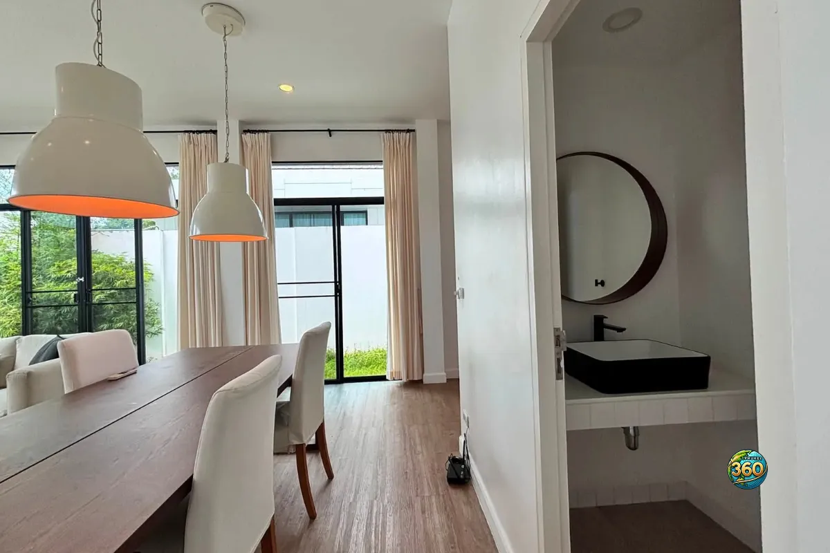 Salle à manger de la maison Mono Plus Palai à Phuket avec table en bois, suspensions blanches, baies vitrées et coin lavabo intérieur