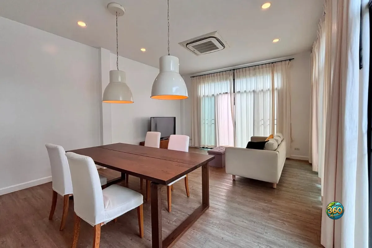 Salle à manger lumineuse de la maison Mono Plus Palai à Phuket avec table en bois, chaises blanches, suspensions et salon en arrière-plan