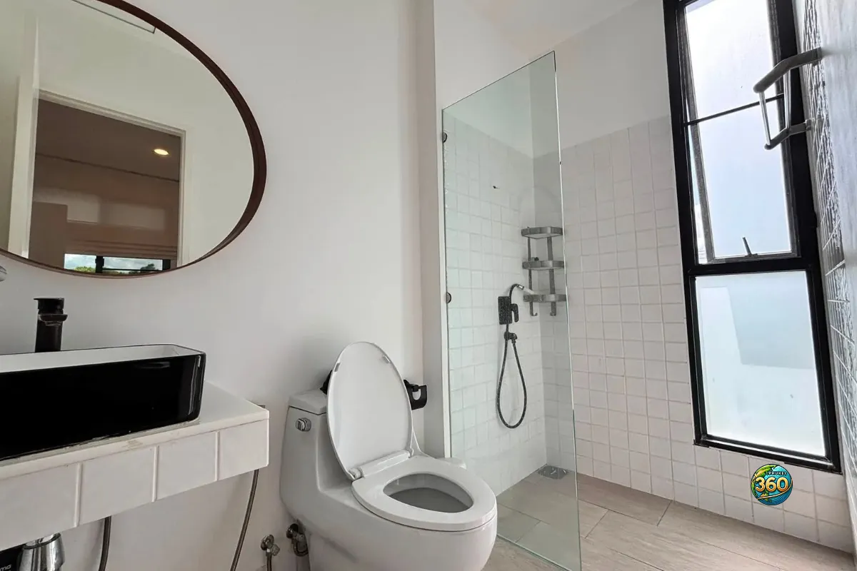 Salle de bain de la maison Mono Plus Palai à Phuket avec douche vitrée, toilettes, lavabo noir, miroir rond et fenêtre verticale