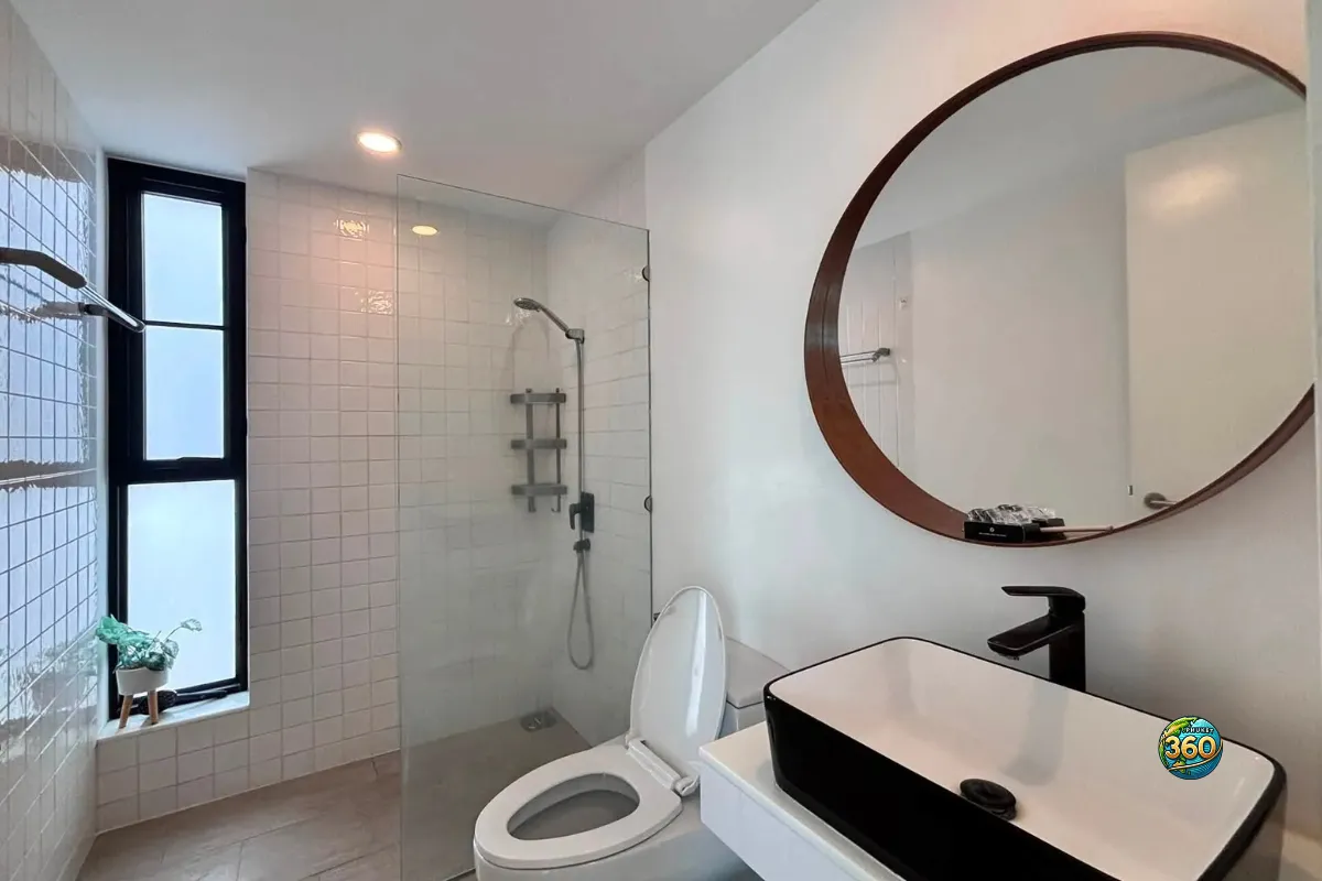 Salle de bain moderne de la maison Mono Plus Palai à Phuket avec douche vitrée, toilettes, lavabo noir et grand miroir rond
