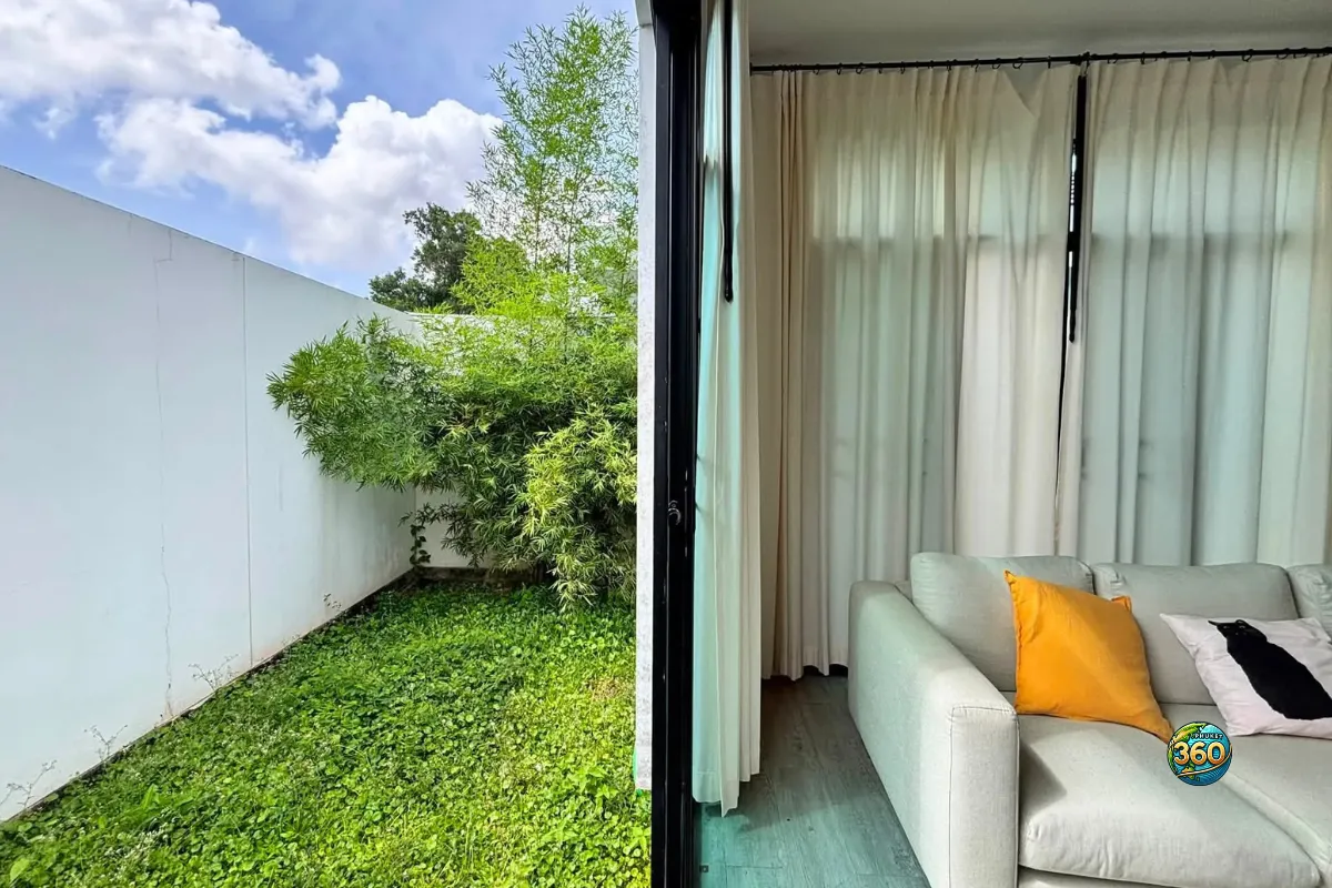 Vue depuis le salon de la maison Mono Plus Palai à Phuket vers le jardin latéral, avec canapé, rideaux et végétation extérieure