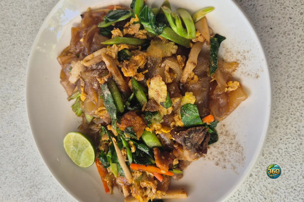 Texte alternatif alt Assiette de pad see ew thaï avec nouilles de riz larges sautées, œuf, légumes verts, poivre et quartier de citron vert